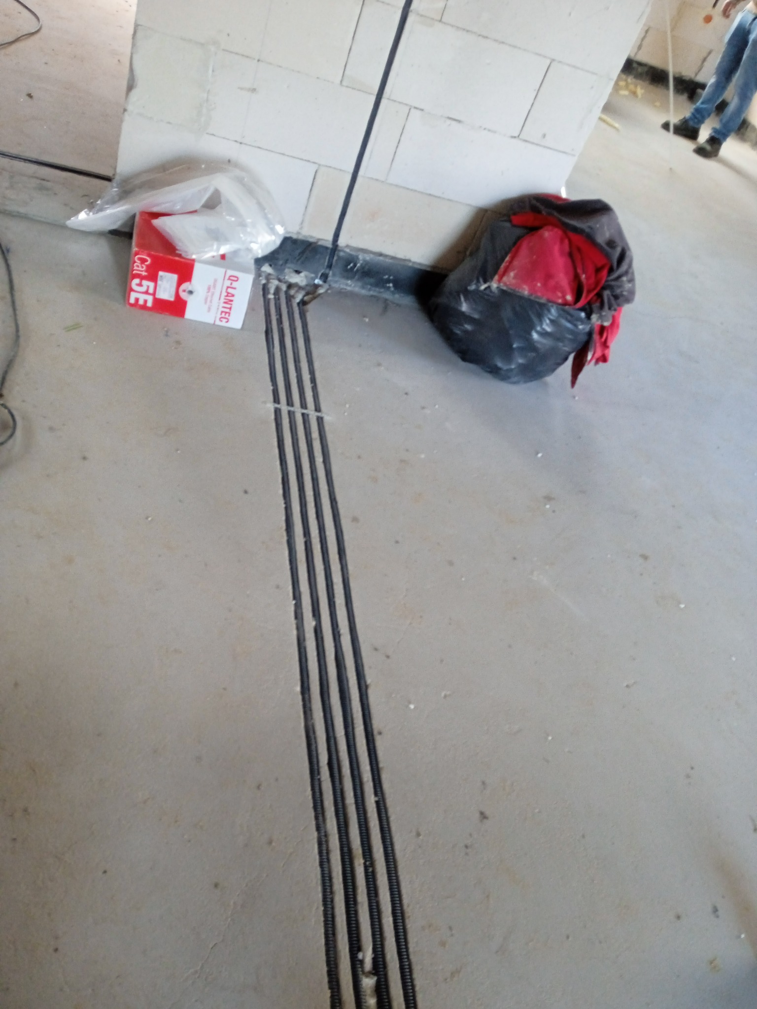 Wykute bruzdy w posadzce pod kable elektryczne, obok puszka instalacyjna i worek z odpadami. Widoczne fragmenty instalacji w ścianie.