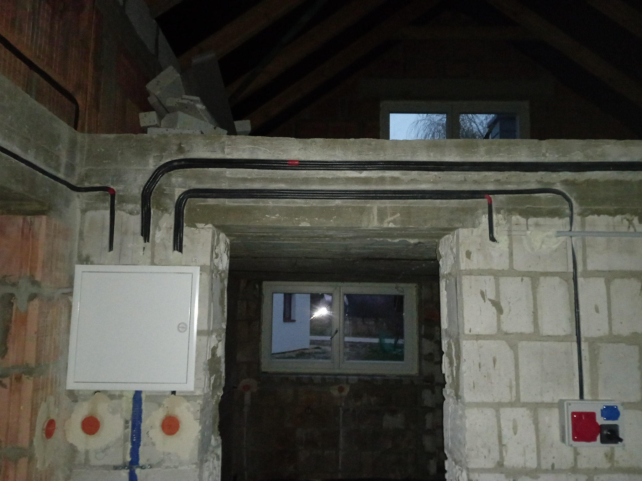 Wnętrze w trakcie prac wykończeniowych z widoczną instalacją elektryczną w peszlach, puszką rozdzielczą i gniazdem siłowym. Surowy stan ścian i stropu.
