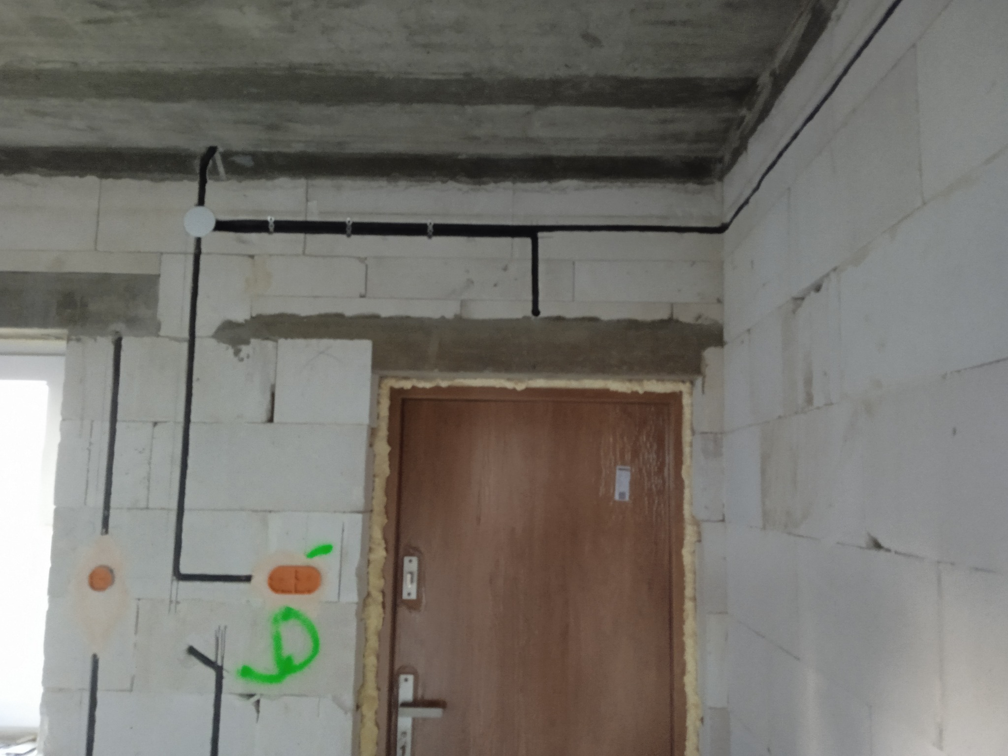 Surowy stan: czarne przewody elektryczne prowadzone po białych ścianach z betonu komórkowego, widoczne puszki instalacyjne i oznaczenia. Drzwi wejściowe w tle.