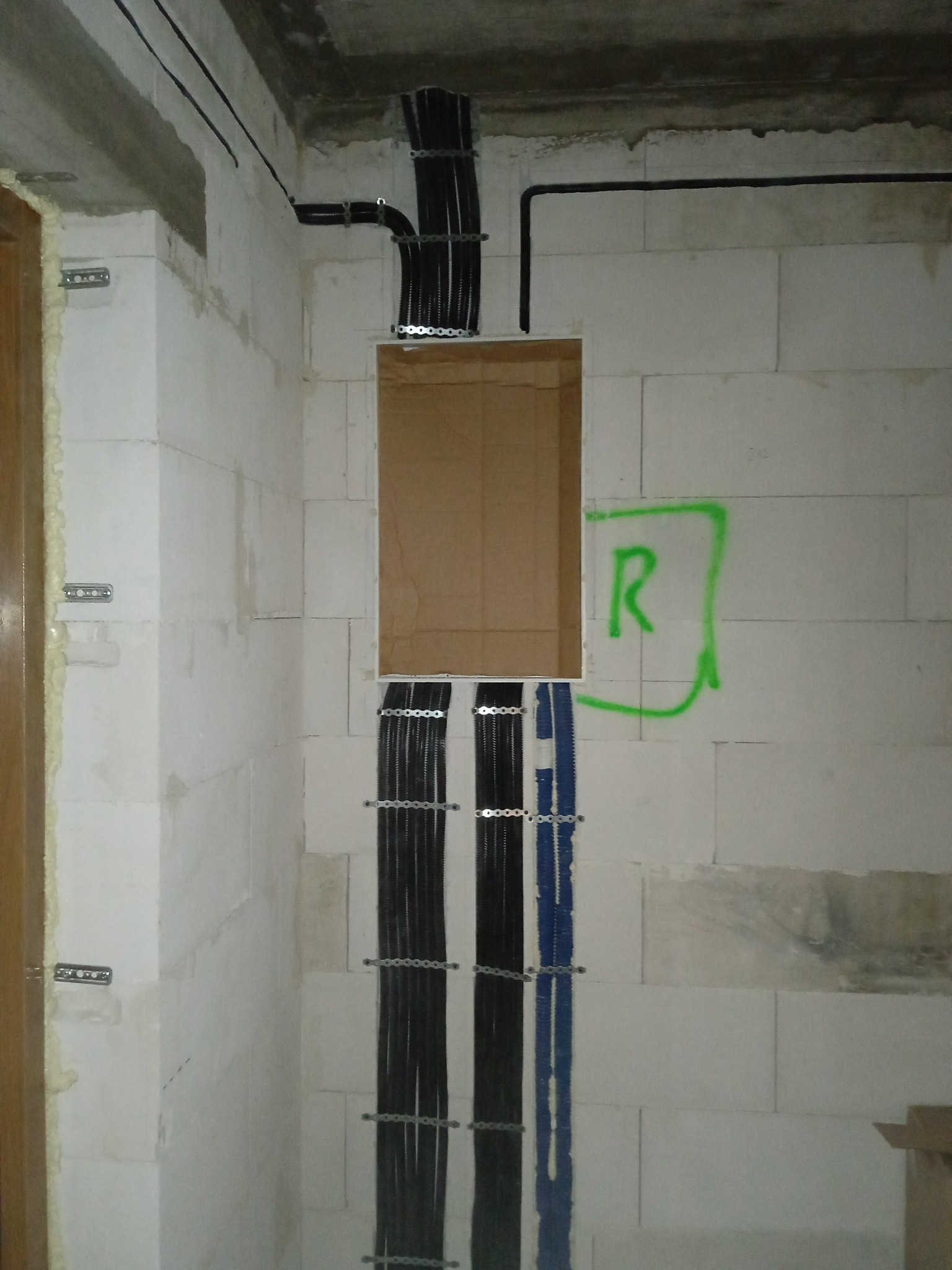 Instalacja elektryczna w trakcie montażu w ścianie z betonu komórkowego. Widoczne kable w peszlach, puszka elektryczna i oznaczenie 'R' na ścianie.
