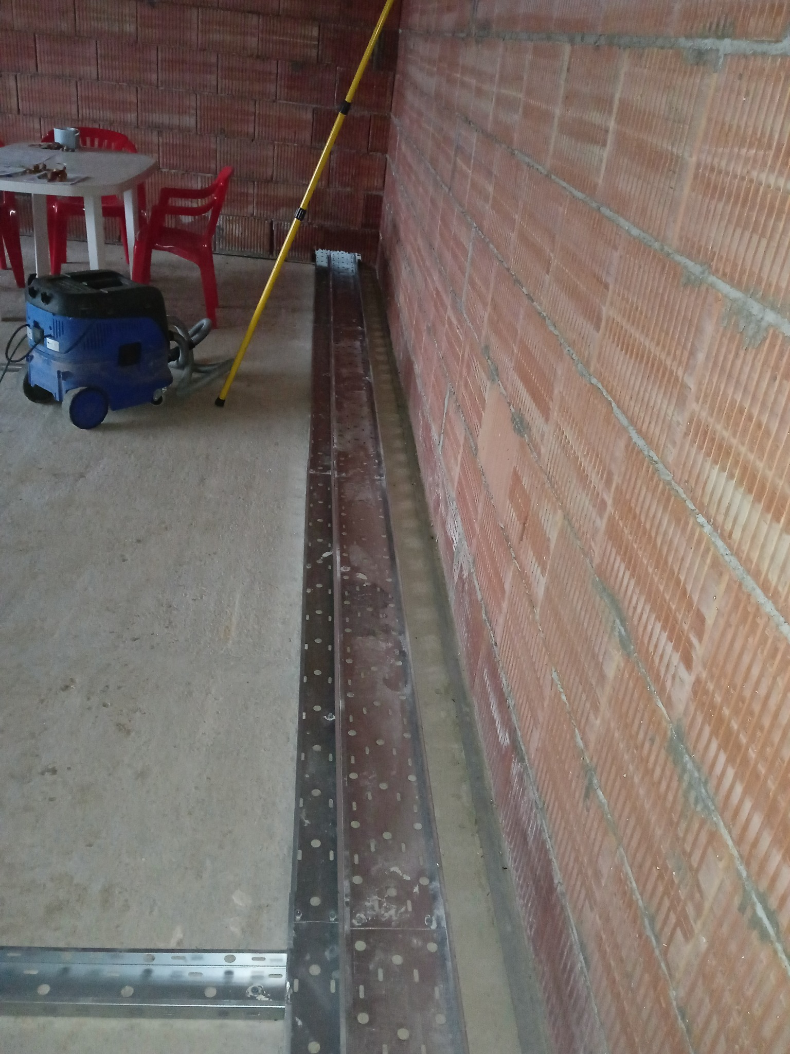 Metalowe korytka kablowe ułożone wzdłuż ceglanej ściany, przygotowane do montażu. W tle widoczny odkurzacz przemysłowy i krzesła.