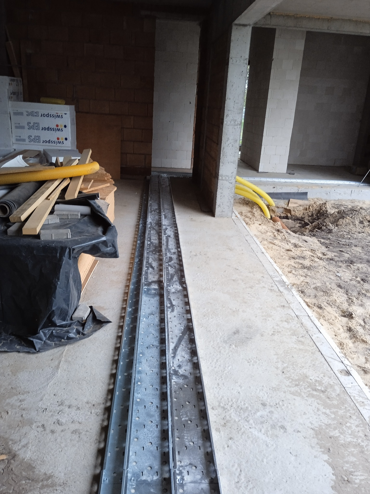 Metalowe korytka kablowe ułożone w surowym wnętrzu budynku w trakcie budowy. Widoczne żółte rury instalacyjne i materiały budowlane w tle. Surowy beton i cegły.