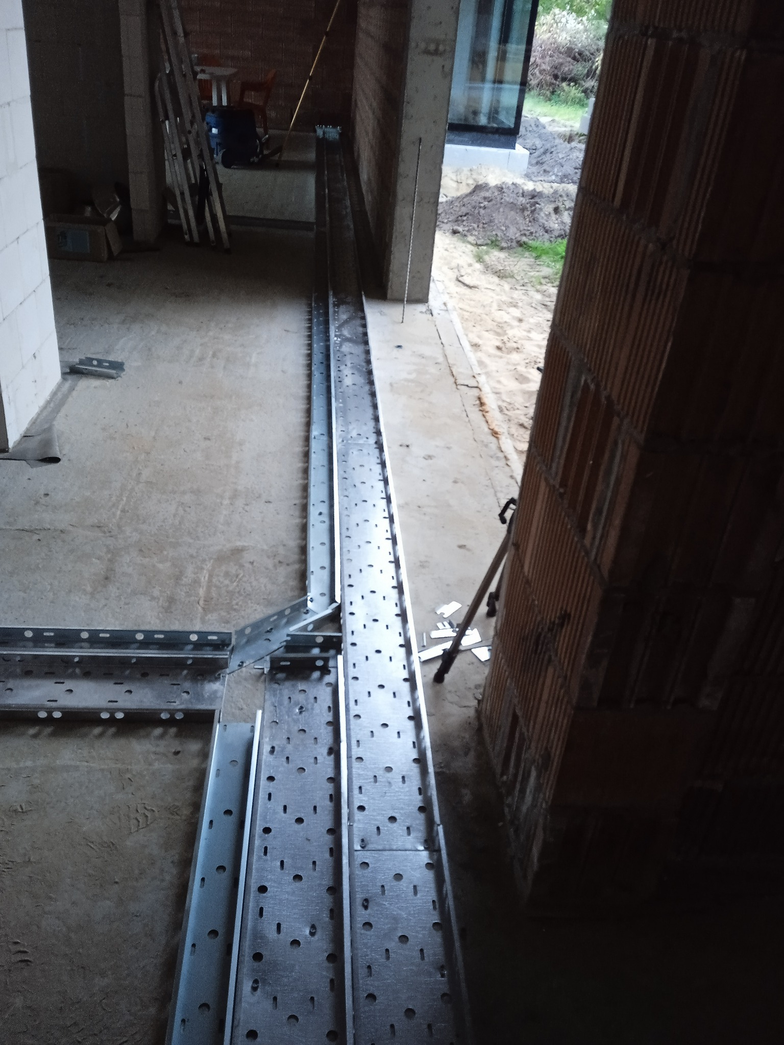 Metalowe korytka kablowe, perforowane, ułożone w surowym wnętrzu budynku w trakcie budowy. Widoczne cegły i betonowe elementy konstrukcyjne.