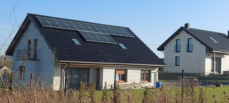 Dom jednorodzinny z zamontowanymi panelami fotowoltaicznymi na dachu z dachówki ceramicznej, widoczne dwa okna dachowe i balkon, w tle drugi dom.