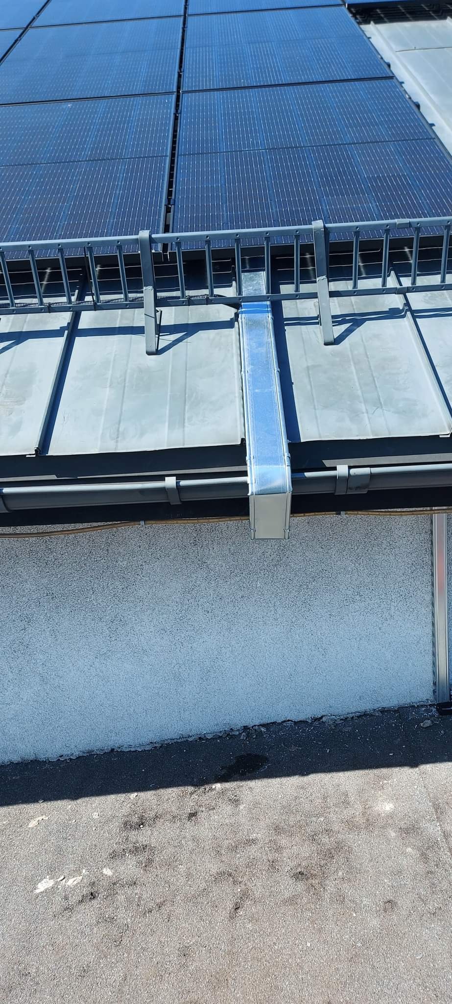 Panel fotowoltaiczny na szarym dachu z metalową konstrukcją wsporczą i rurą spustową, widoczne elementy elewacji budynku.