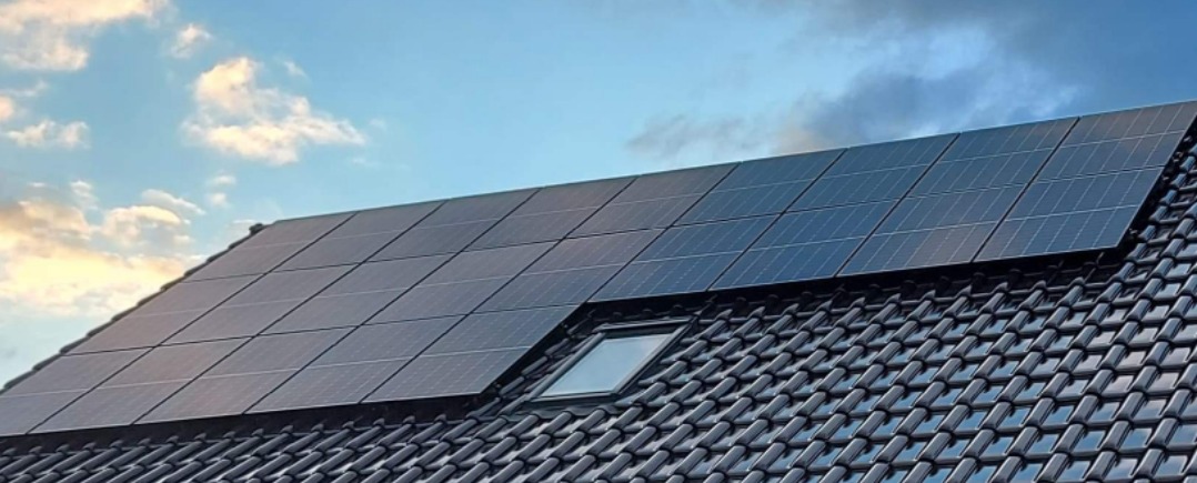 Panel fotowoltaiczny na dachu z dachówką w kolorze antracytowym, widoczny lukarna, błękitne niebo z chmurami w tle.