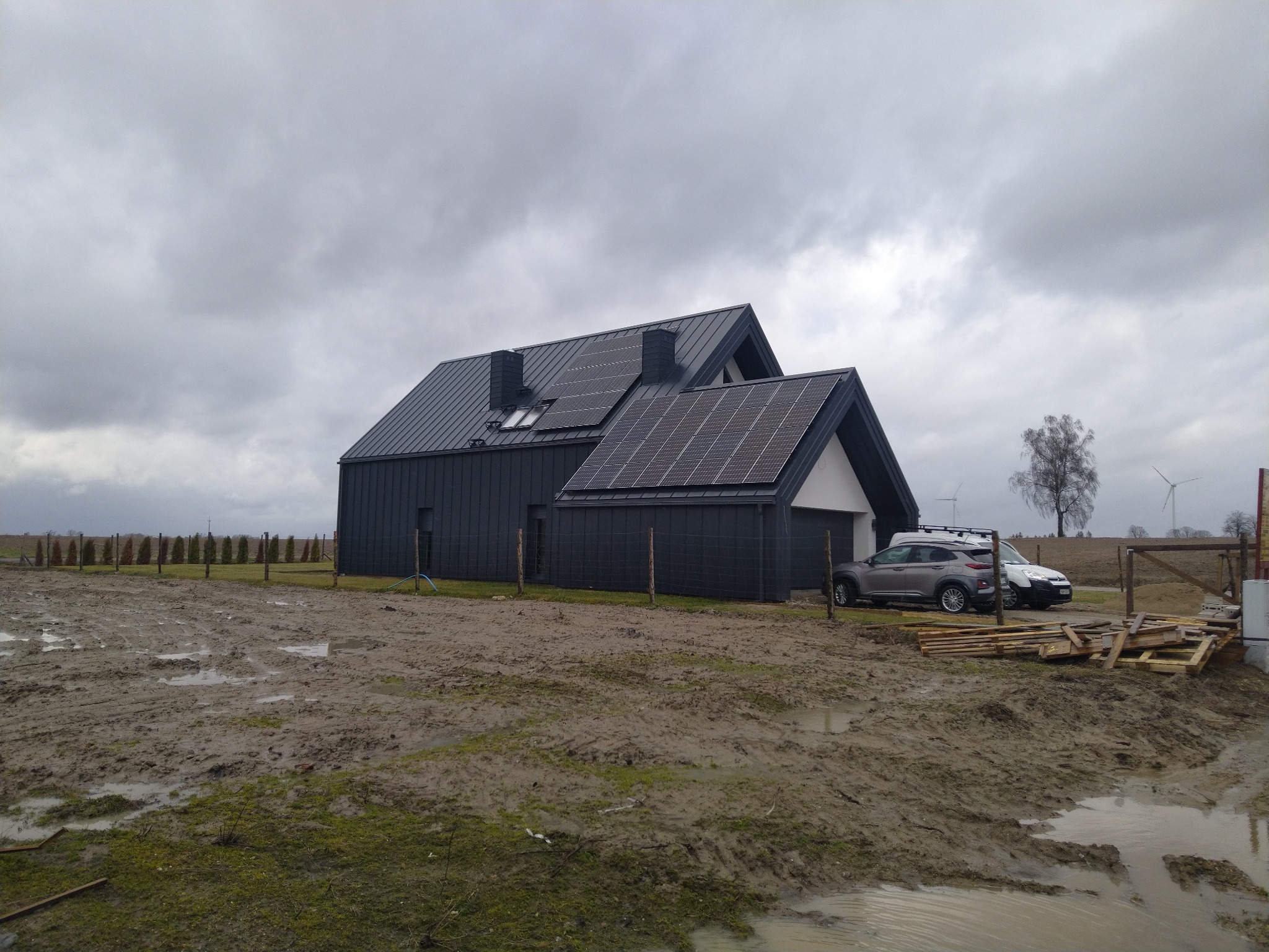 Nowoczesny dom z ciemną elewacją i panelami słonecznymi na dachu w pochmurny dzień, teren wokół w trakcie zagospodarowywania, widoczne dwa samochody i elementy drewniane.