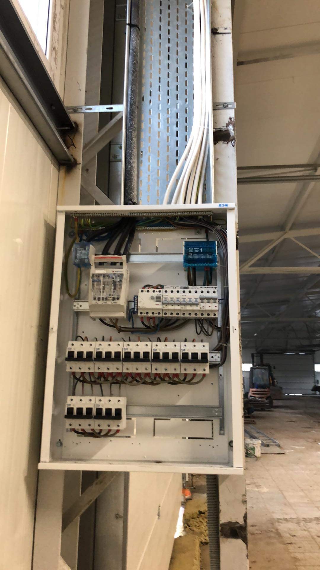 Rozdzielnica elektryczna w metalowej obudowie, z widocznymi bezpiecznikami, przewodami i listwami zaciskowymi, zamontowana na ścianie w hali przemysłowej, z widoczną instalacją kablową w korycie...