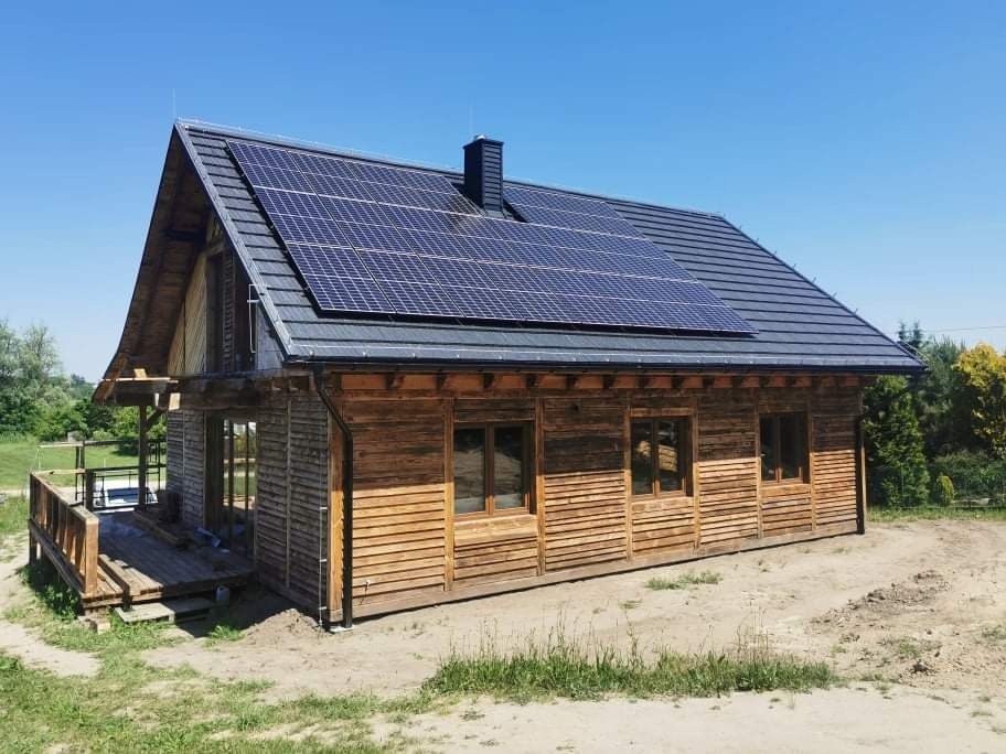 Drewniany dom z ciemną dachówką i panelami słonecznymi na dachu, widok z boku w słoneczny dzień, na trawniku