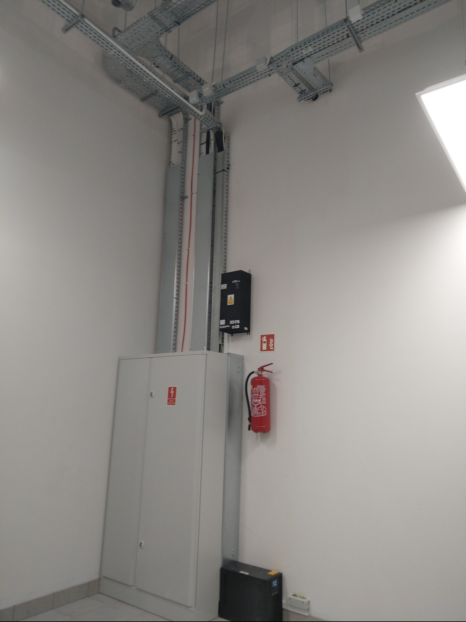 Szara szafa elektryczna z oznaczeniem, czarna skrzynka, gaśnica i koryta kablowe pod sufitem w jasnym pomieszczeniu. Widok z dołu.