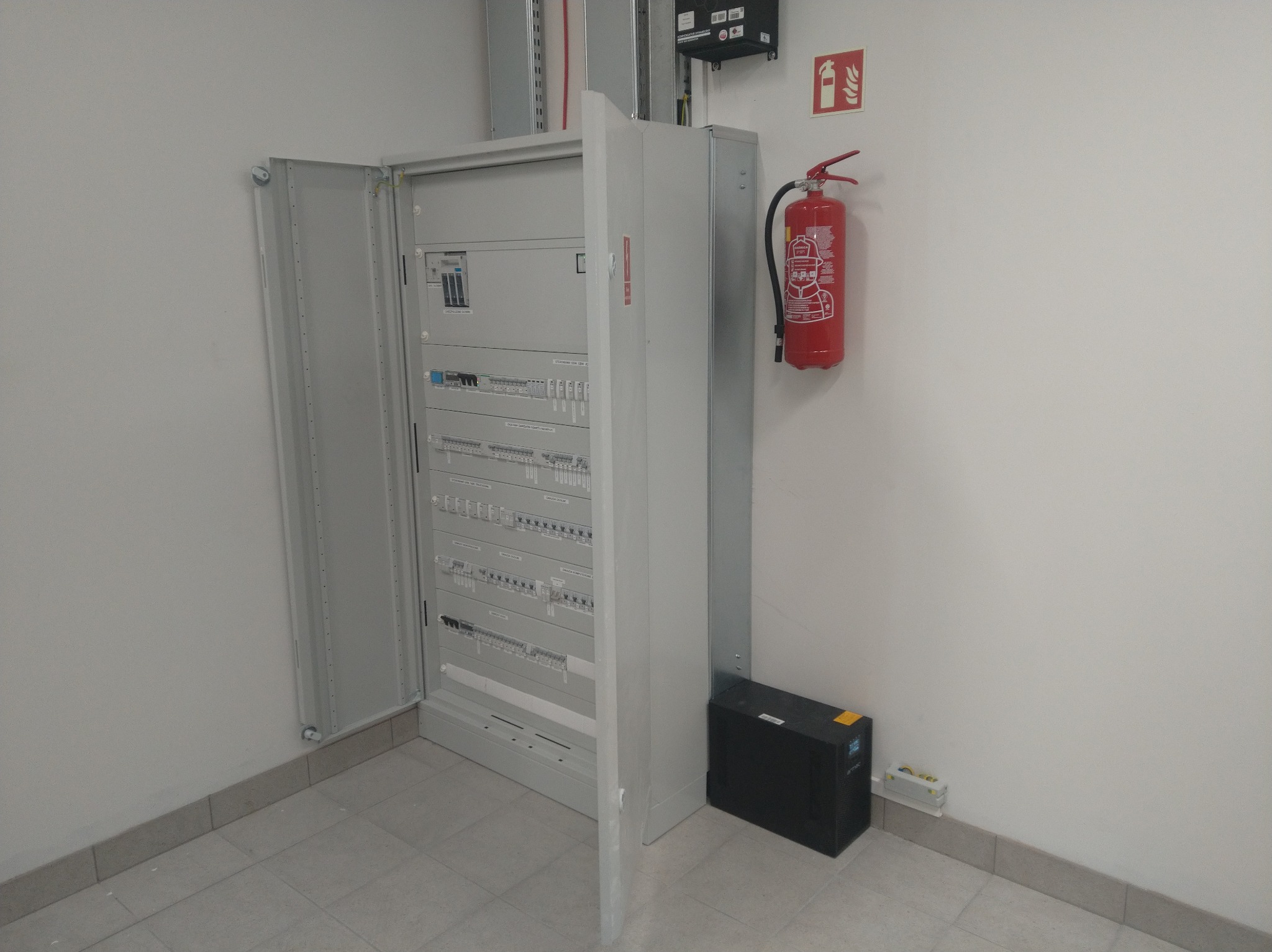 Otwarta szafa elektryczna z widocznymi bezpiecznikami i okablowaniem, obok gaśnica i zasilacz awaryjny UPS. Widok na instalację w pomieszczeniu.