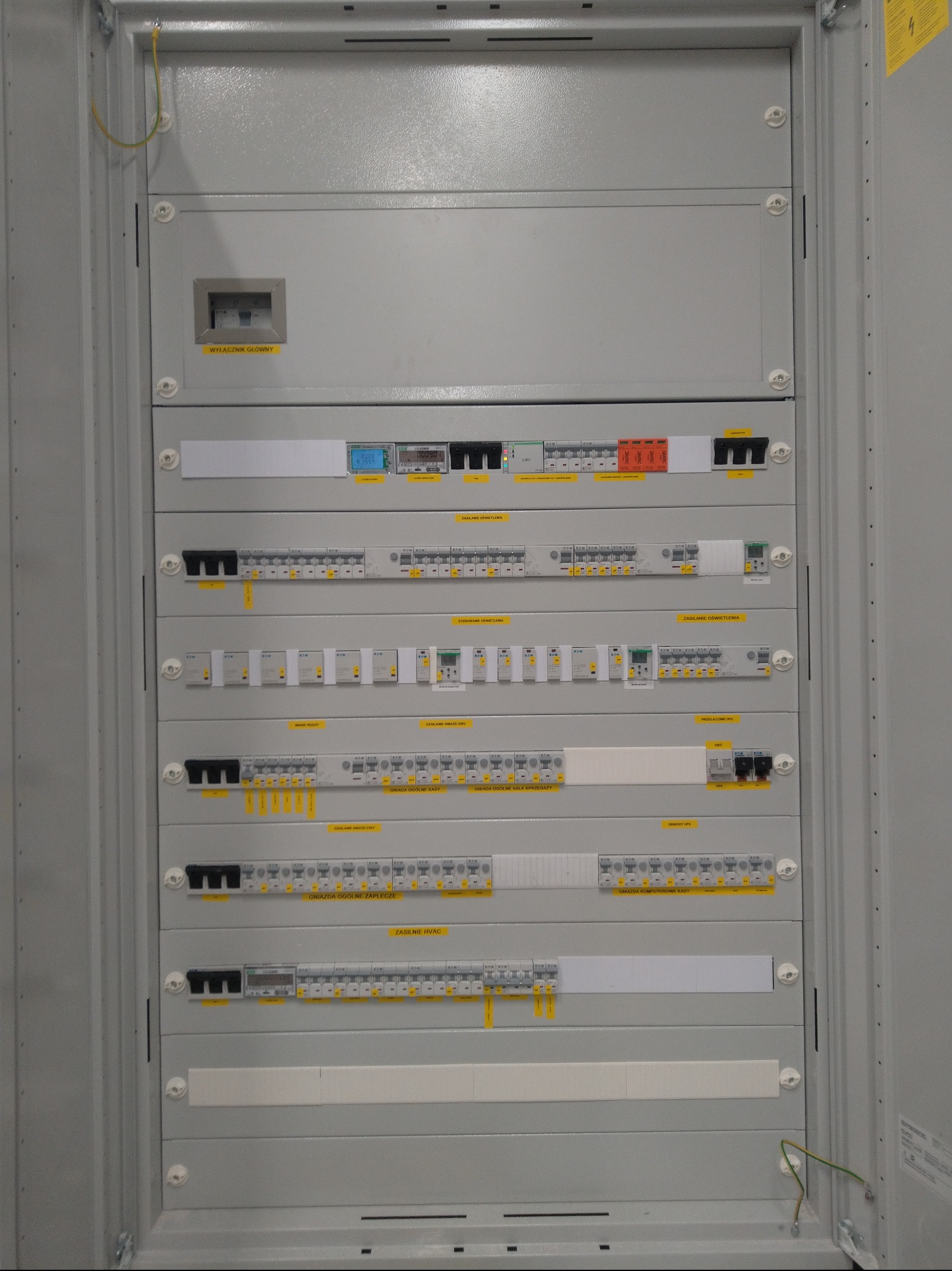 Szara szafa elektryczna z bezpiecznikami, wyłącznikami i okablowaniem. Widoczne żółte etykiety z opisami obwodów. Instalacja wewnątrz szafy uporządkowana.