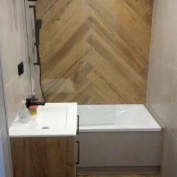 Wykończona łazienka z drewnopodobną glazurą ułożoną w jodełkę na podłodze i ścianie nad wanną, biała wanna z zabudową, umywalka nablatowa z szafką z frontami w kolorze drewna, czarna bateria...