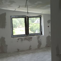 Db-Rem - Wnętrze pokoju w stanie surowym po nałożeniu gładzi, widoczne ślady po szpachlowaniu na ścianach i suficie, okno z widokiem na zieleń, wystające przewody elektryczne ze ściany i sufitu.