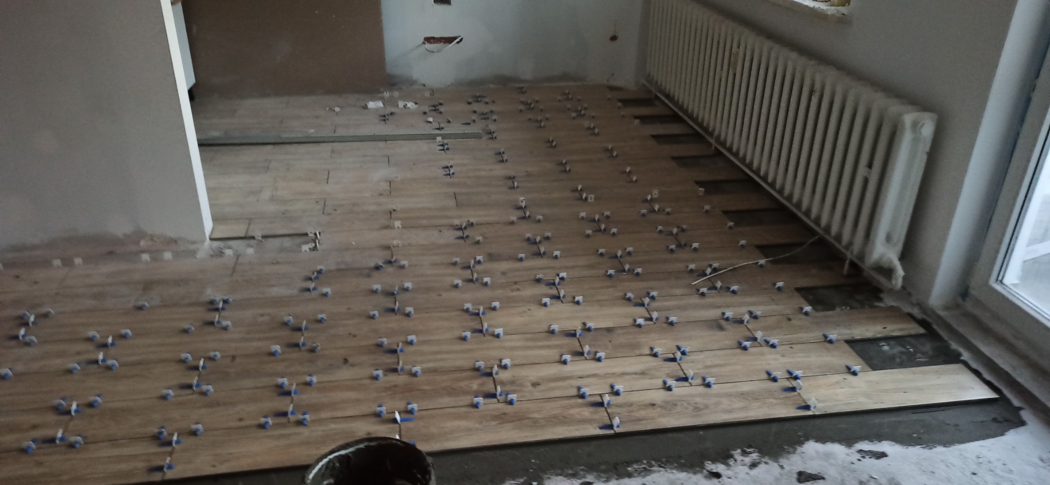 Układanie podłogi z płytek imitujących drewno, z użyciem niebieskich krzyżyków dystansowych; widoczny grzejnik i okno w tle.