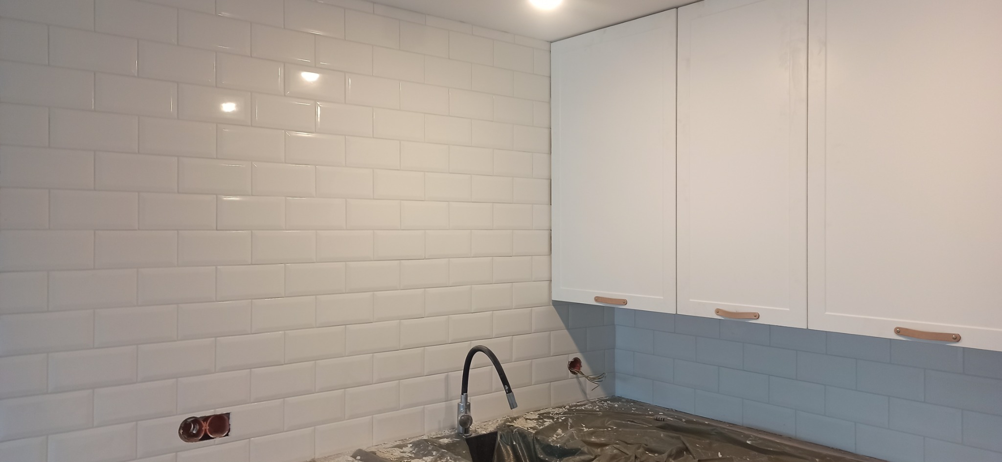 Białe, błyszczące płytki ułożone w cegiełkę na ścianie kuchni, widoczne białe szafki z jasnobrązowymi uchwytami, czarny kran, instalacja elektryczna bez osprzętu w ścianie, zabezpieczony blat.