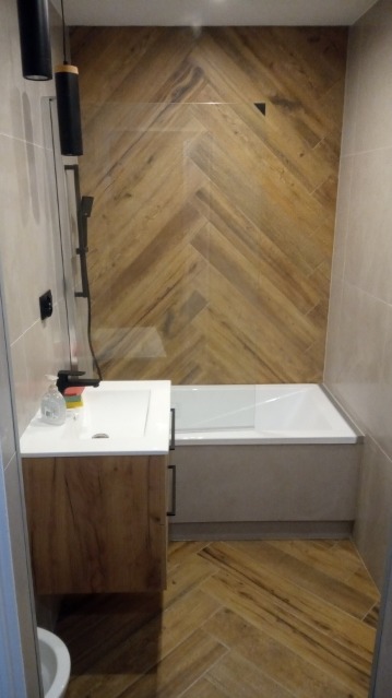 Wykończona łazienka z drewnopodobną glazurą ułożoną w jodełkę na podłodze i ścianie nad wanną, biała wanna z zabudową, umywalka nablatowa z szafką z frontami w kolorze drewna, czarna bateria...