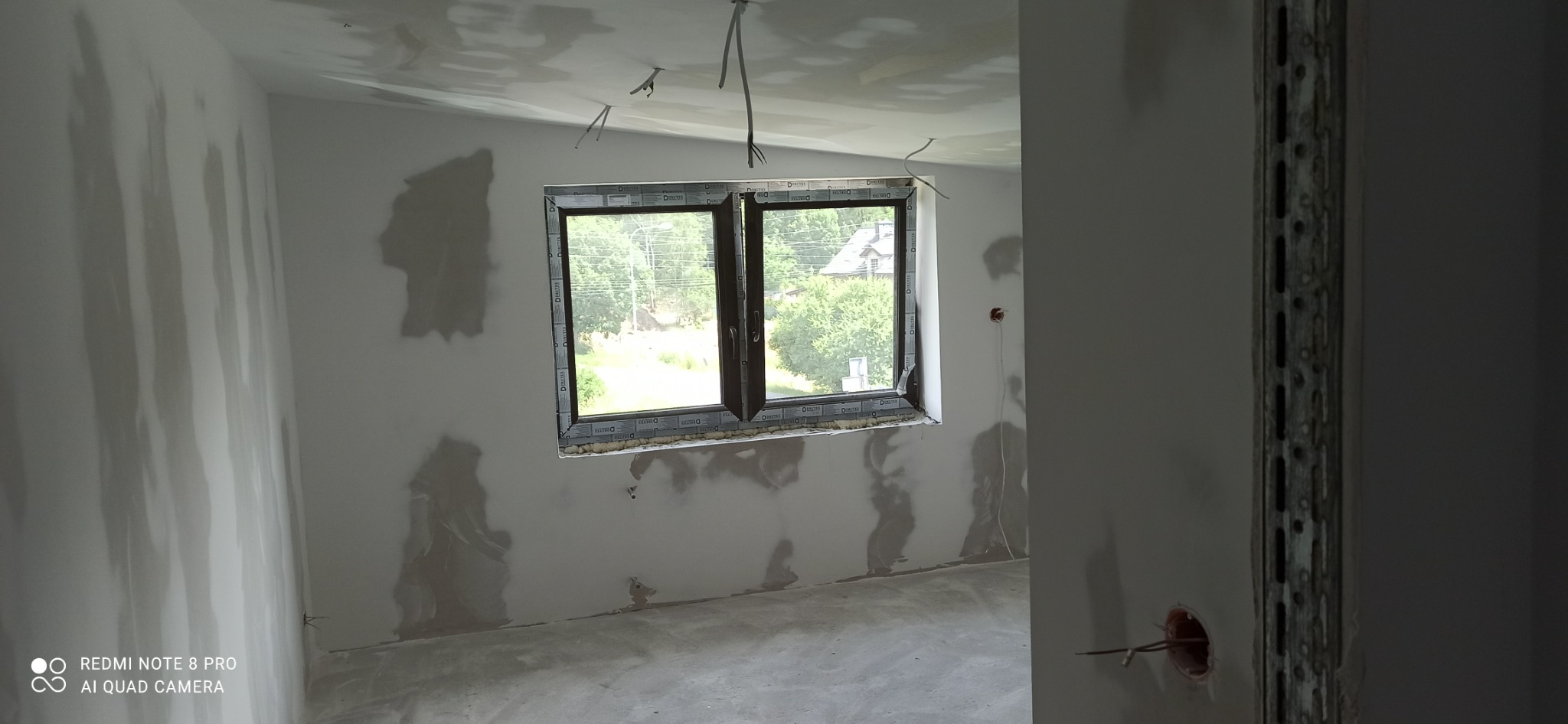 Wnętrze pokoju w stanie surowym po nałożeniu gładzi, widoczne ślady po szpachlowaniu na ścianach i suficie, okno z widokiem na zieleń, wystające przewody elektryczne ze ściany i sufitu.