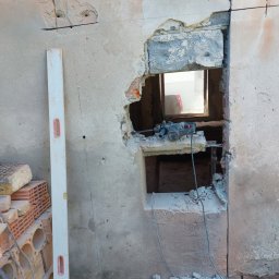 MAR TOM - Proces wykuwania otworu w ścianie z cegły, widoczny poziomica przy ścianie, gruz i cegły na podłodze, w otworze umieszczona wiertarka udarowa.