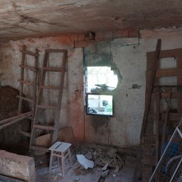 MAR TOM - Wnętrze starego, zniszczonego pomieszczenia z fragmentami gruzu, drewnianymi drabinami opartymi o ściany i widokiem na zewnątrz przez wybite okno.