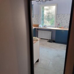 MAR TOM - Widok przez drzwi na wyremontowaną kuchnię w Bielsku-Białej: jasne płytki podłogowe, niebieskie szafki, biały grzejnik i ściany pomalowane na fioletowo z wzorzystą tapetą nad blatem.