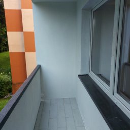 MAR TOM - Wykończony balkon z szarymi płytkami podłogowymi, świeżo pomalowanymi ścianami na szaro i oknem z czarnym parapetem, widok na zewnątrz na zieleń i fragment budynku w pomarańczowo-białą kratę.