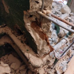 MAR TOM - Uszkodzony fragment ściany z odsłoniętym zbrojeniem, resztki starej, zielonej farby i gruz budowlany.