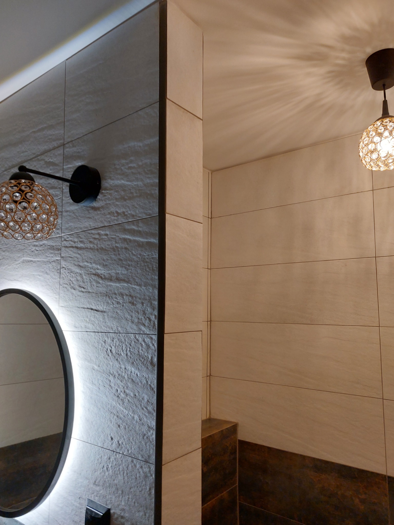 Nowoczesna łazienka z szarymi płytkami imitującymi kamień, okrągłym lustrem podświetlanym LED, kryształową lampą ścienną i sufitową, oraz brązowymi akcentami w dolnej części ściany.