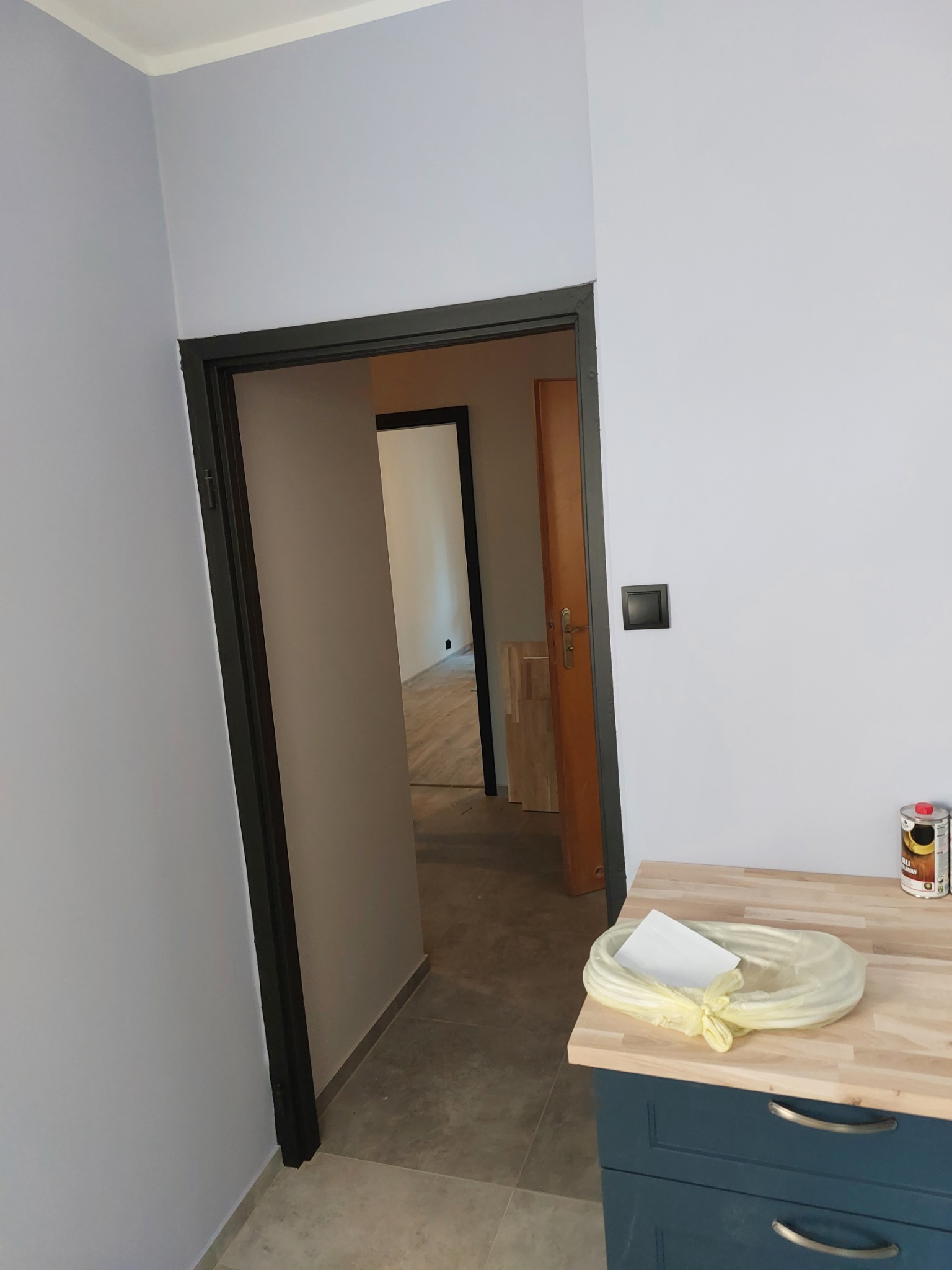 Świeżo pomalowane, jasnoniebieskie ściany z czarną ościeżnicą, widok na sąsiednie pomieszczenie z wykończoną podłogą, na pierwszym planie blat z akcesoriami malarskimi i puszką farby.