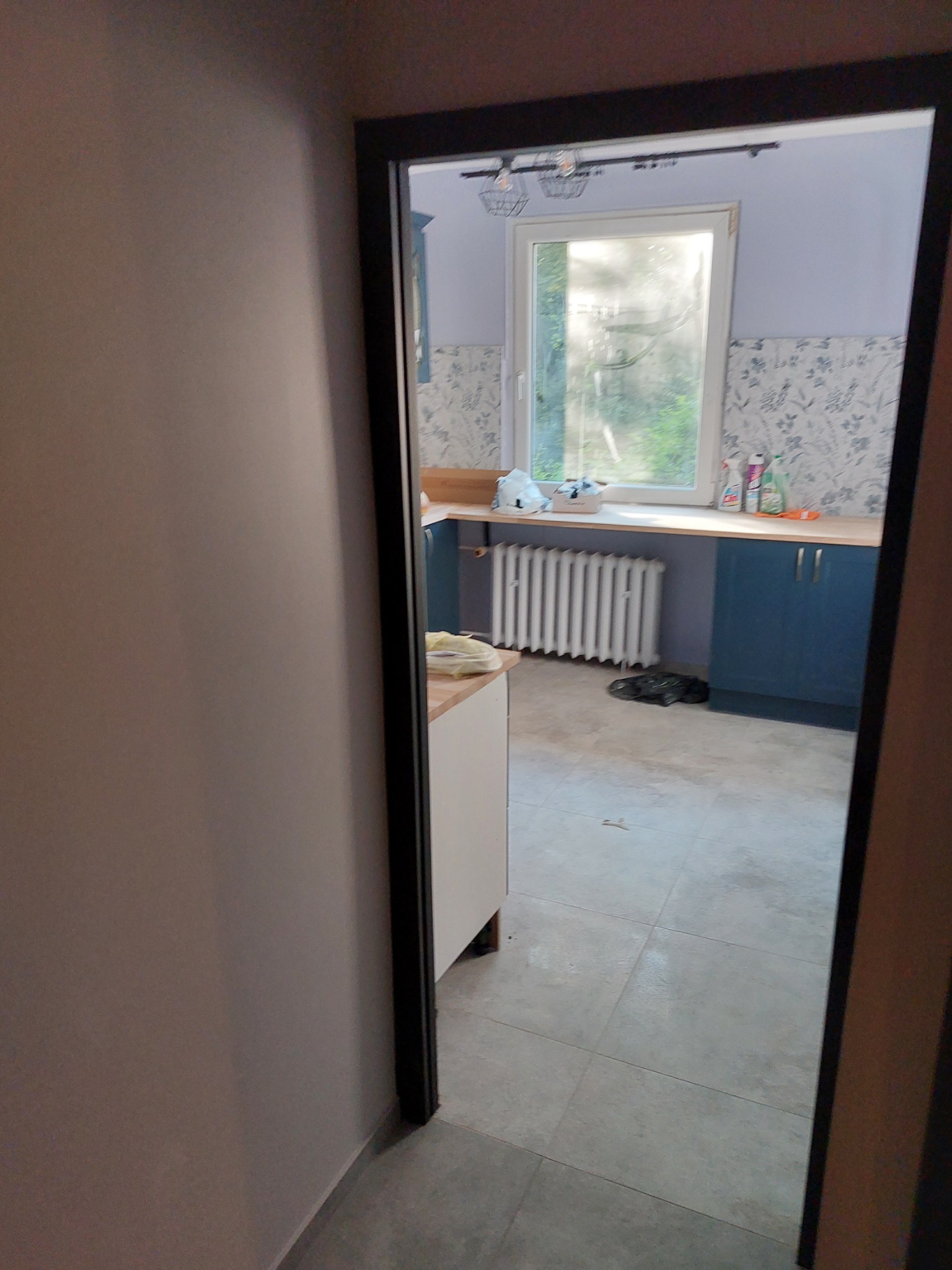 Widok przez drzwi na wyremontowaną kuchnię w Bielsku-Białej: jasne płytki podłogowe, niebieskie szafki, biały grzejnik i ściany pomalowane na fioletowo z wzorzystą tapetą nad blatem.