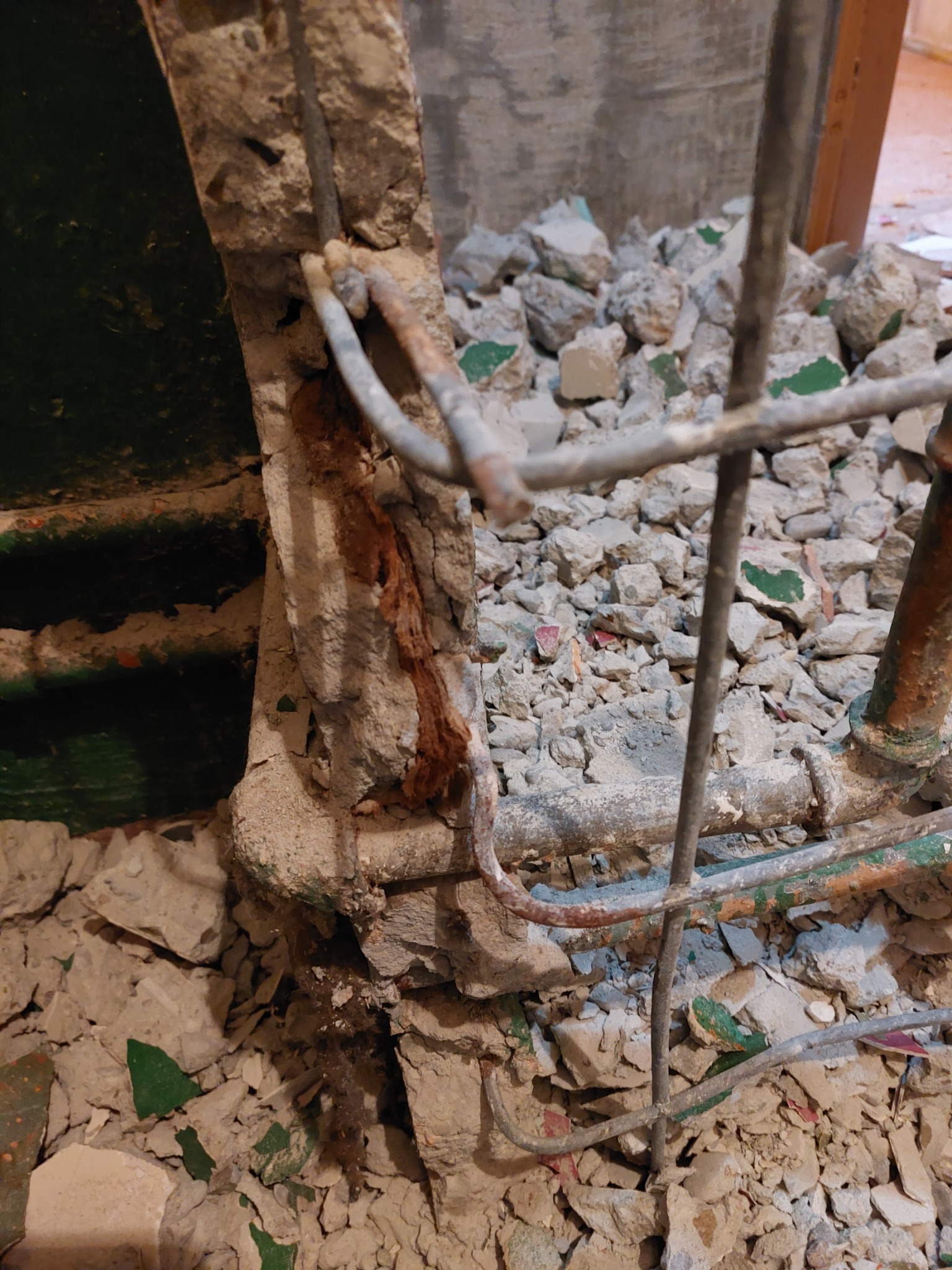Fragment ściany z odsłoniętym zbrojeniem i gruzem po skuciu tynku, widoczne fragmenty starej zielonej farby.