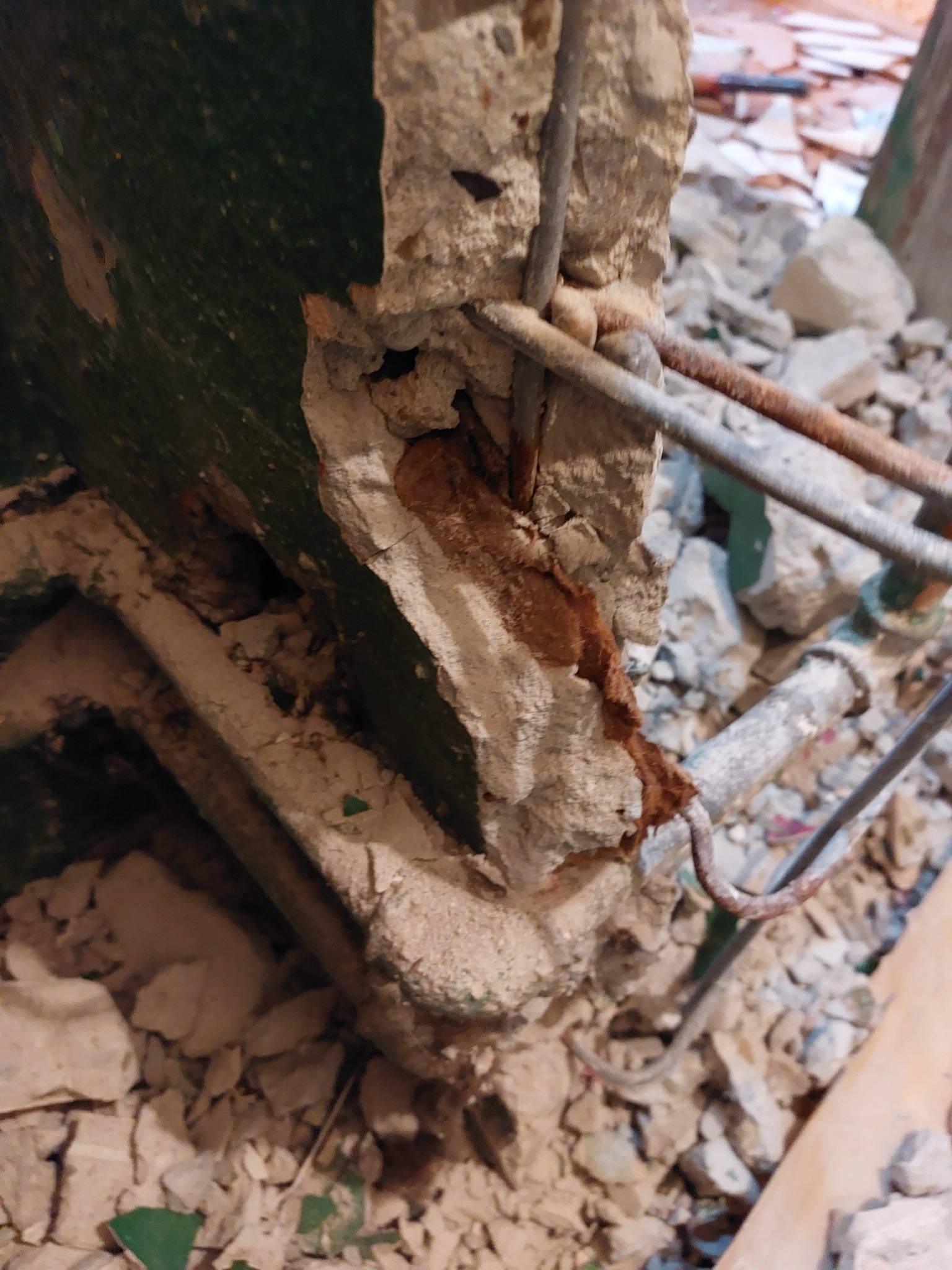 Uszkodzony fragment ściany z odsłoniętym zbrojeniem, resztki starej, zielonej farby i gruz budowlany.