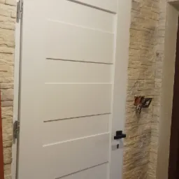 Białe drzwi wewnętrzne z poziomymi żłobieniami, zamontowane na tle ściany wykończonej dekoracyjną okładziną imitującą kamień, z widoczną puszką elektryczną bez osprzętu.