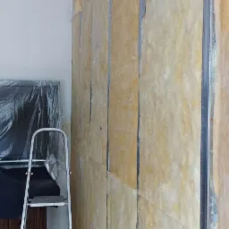 Ściana z zamontowaną wełną mineralną pomiędzy metalowymi profilami, widoczna drabina aluminiowa i folia ochronna w tle.