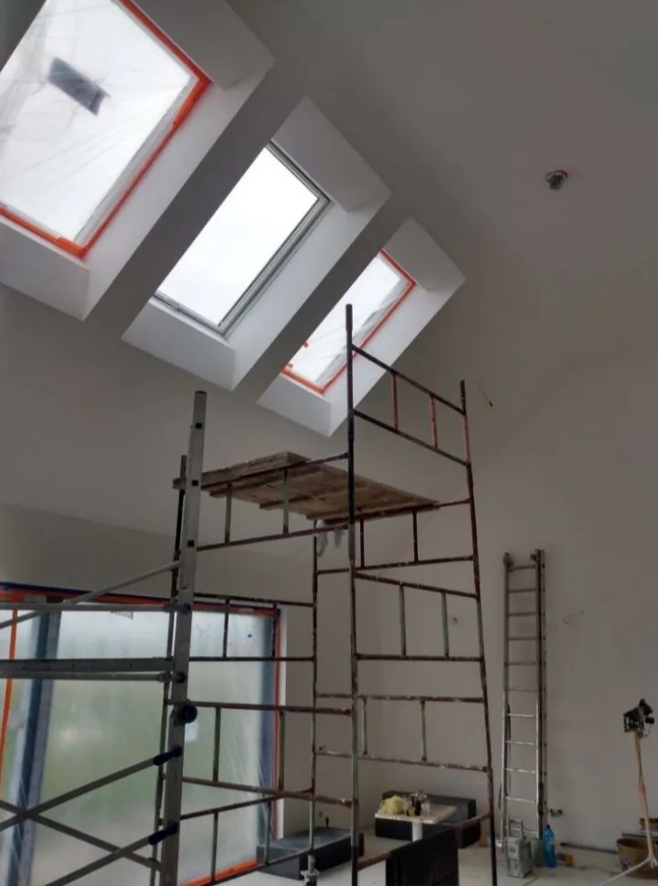 Wnętrze poddasza ze świeżo pomalowanymi ścianami, widoczne trzy okna dachowe, rusztowanie i drabina oparta o ścianę, przygotowane do dalszych prac wykończeniowych.