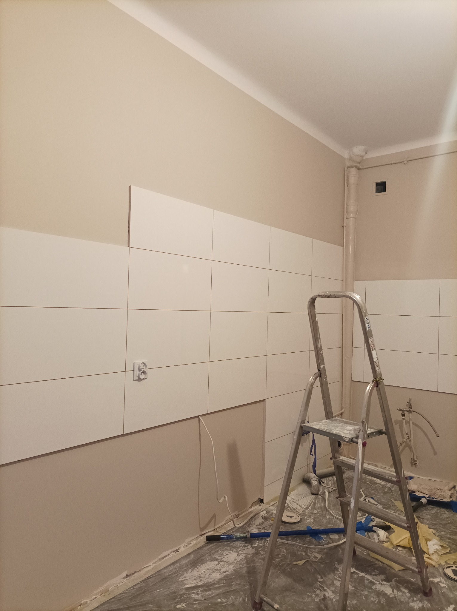 Wnętrze w trakcie układania białych płytek ściennych, widoczna aluminiowa drabina, częściowo zakryte podłoże folią ochronną, gniazdko elektryczne i rura pionowa. Jedna płytka brakuje.
