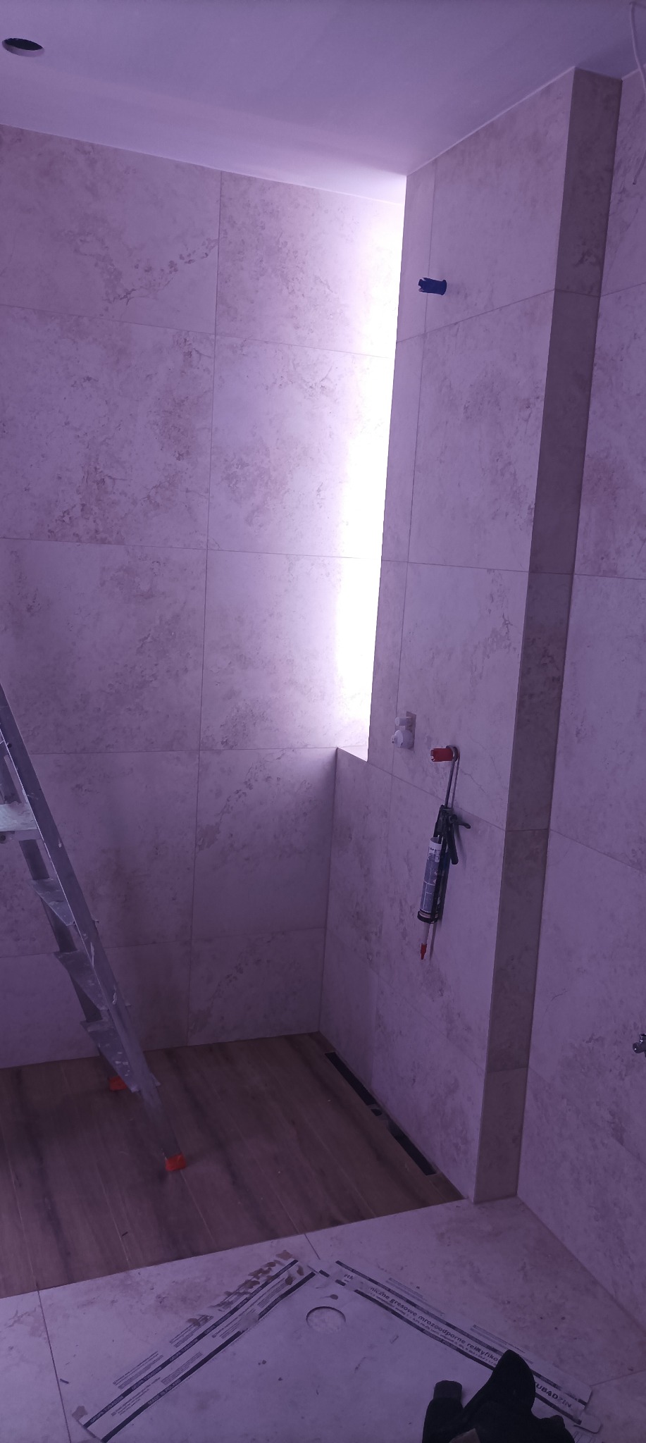 Wnętrze łazienki w trakcie wykańczania, widoczne płytki imitujące marmur na ścianach, drewnopodobna podłoga, drabina malarska i aplikator do silikonu z kartuszem, oświetlenie punktowe w suficie.