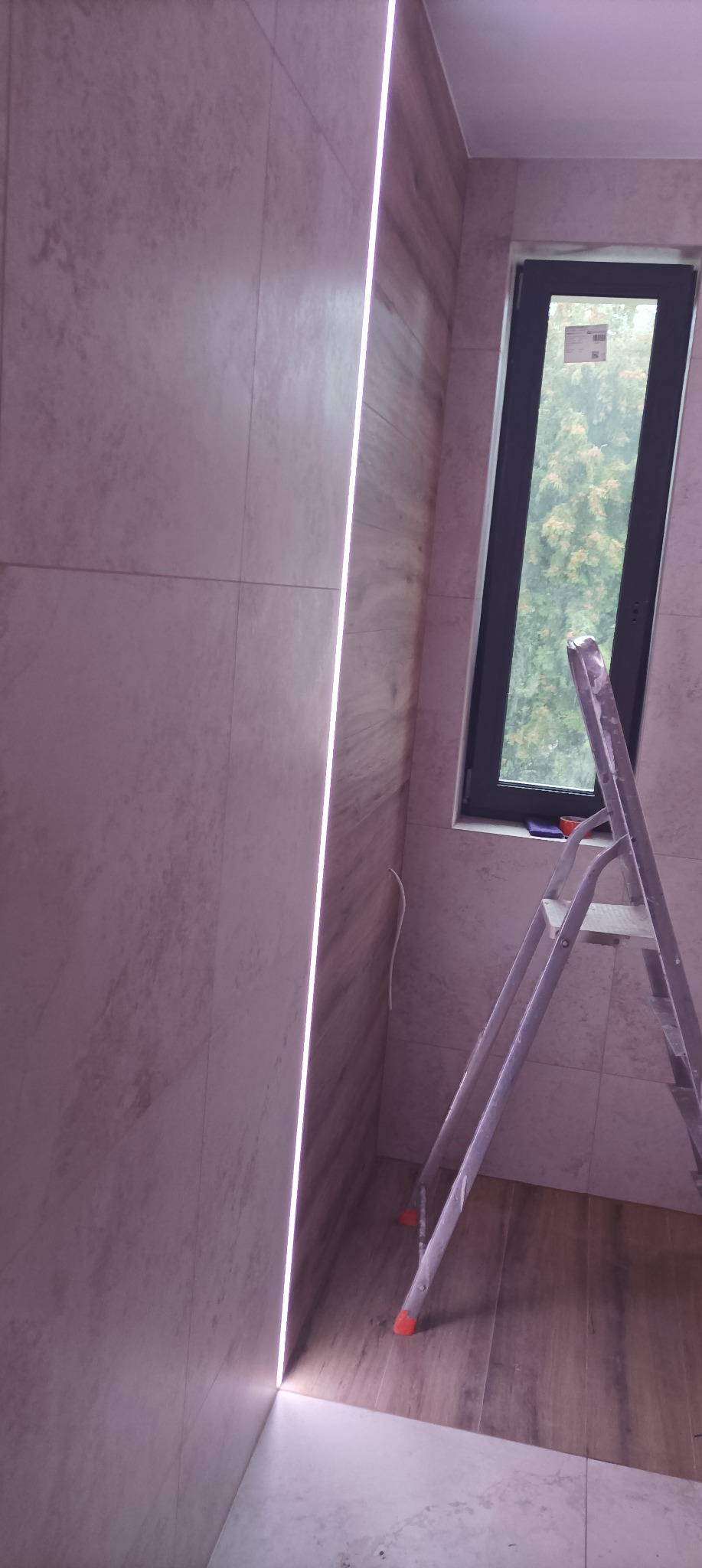 Ściana wyłożona płytkami w łazience z wbudowanym oświetleniem LED, widoczna drabina aluminiowa i okno z widokiem na zieleń.