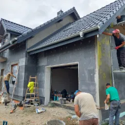 Ekipa budowlana podczas prac elewacyjnych na dwukondygnacyjnym domu z szarych bloczków, widoczne rusztowania, drabiny, wiadra i narzędzia, fragment dachu z ciemnej dachówki.