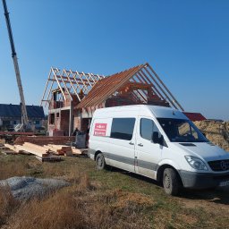 Jar-Mich s.c - Budowa domu z czerwonych bloczków z widoczną drewnianą więźbą dachową, obok biały bus Mercedes-Benz firmy JAR MICH, wysięgnik dźwigu i stosy drewna pod błękitnym niebem.