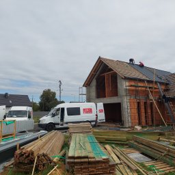Jar-Mich s.c - Budowa domu z pustaków ceramicznych, gdzie dwóch pracowników montuje ciemny dach. Na pierwszym planie stosy drewnianych łat i paneli dachowych, w tle białe busy budowlane.