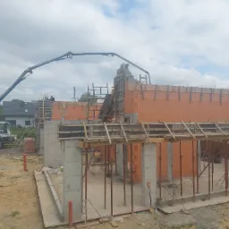 Budowa domu z czerwonej cegły ceramicznej, widoczne betonowe słupy, szalunki drewniane, pompa do betonu w tle i pracownik na rusztowaniu.