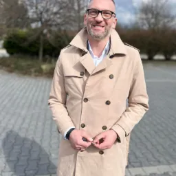 Uśmiechnięty mężczyzna w okularach i beżowym płaszczu, stojący na zewnątrz na tle drzew i pochmurnego nieba.