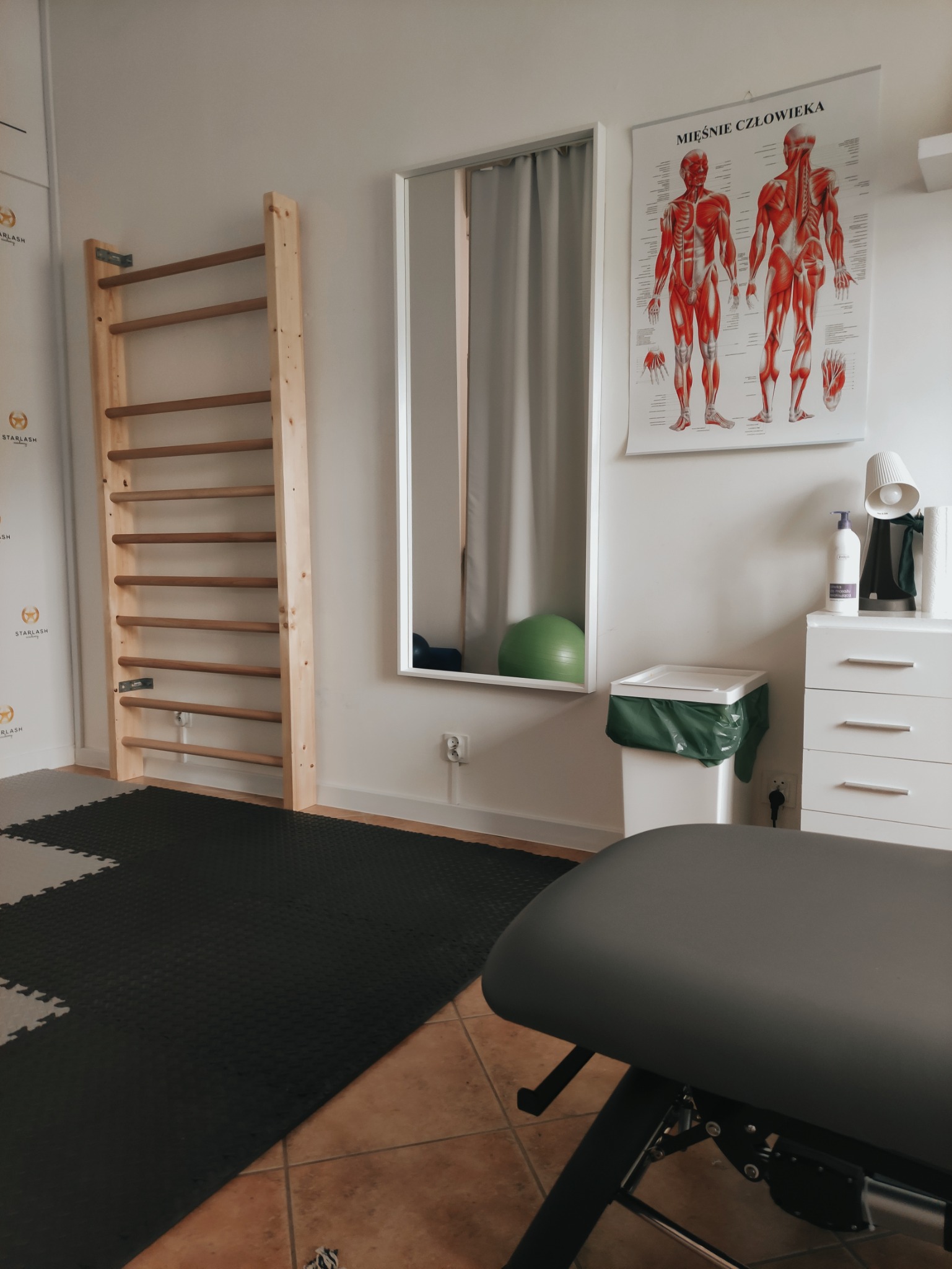 Gabinet z drabinką rehabilitacyjną, stołem do masażu, plakatem anatomicznym i piłką gimnastyczną odbijającą się w lustrze.