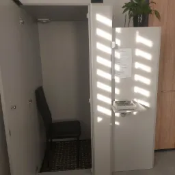 Wyciszony pokój do badań audiometrycznych z krzesłem i aparaturą pomiarową na półce, oświetlony światłem dziennym wpadającym przez żaluzje.