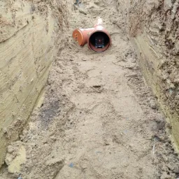 Pomarańczowa rura kanalizacyjna z laserowym niwelatorem umieszczona w wykopie z piaskową podsypką; w tle widoczna łopata.