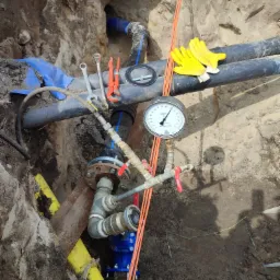 Instalacja wodociągowa w wykopie z widocznym manometrem, żółtymi rękawicami roboczymi, kluczami i fragmentem niebieskiej rury w tle.