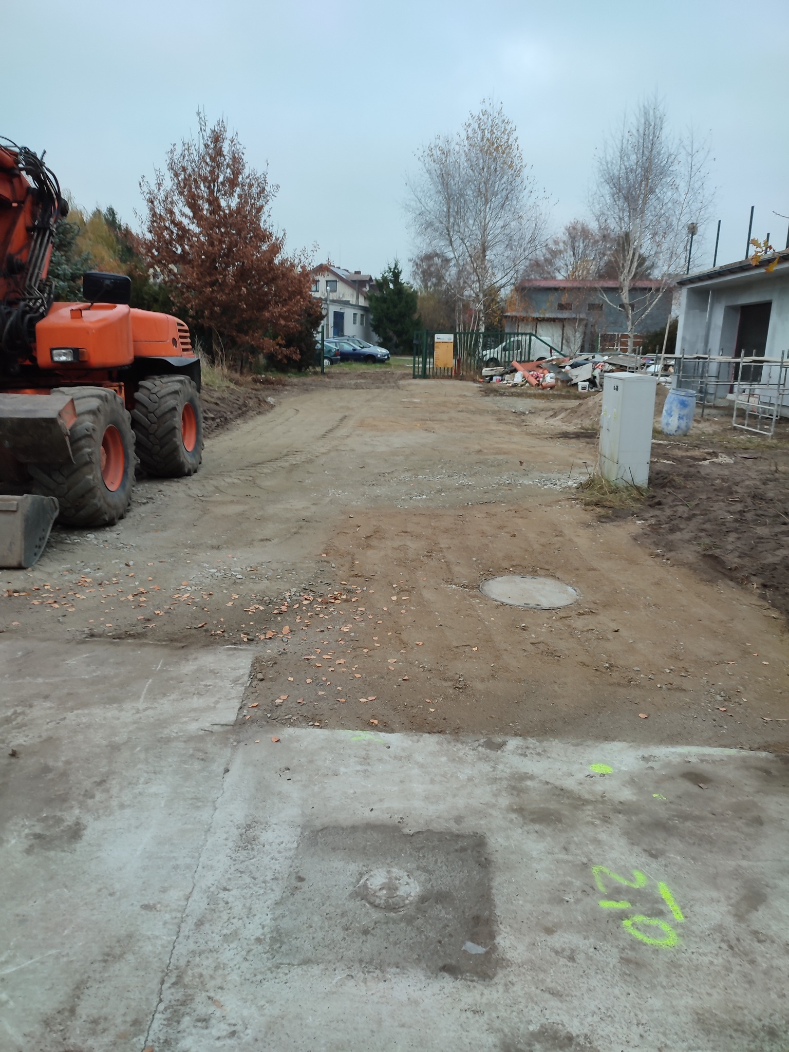 Pomarańczowa koparka kołowa na placu budowy, widoczne ślady opon na ziemi, w tle budynek w trakcie budowy i drzewa bez liści.