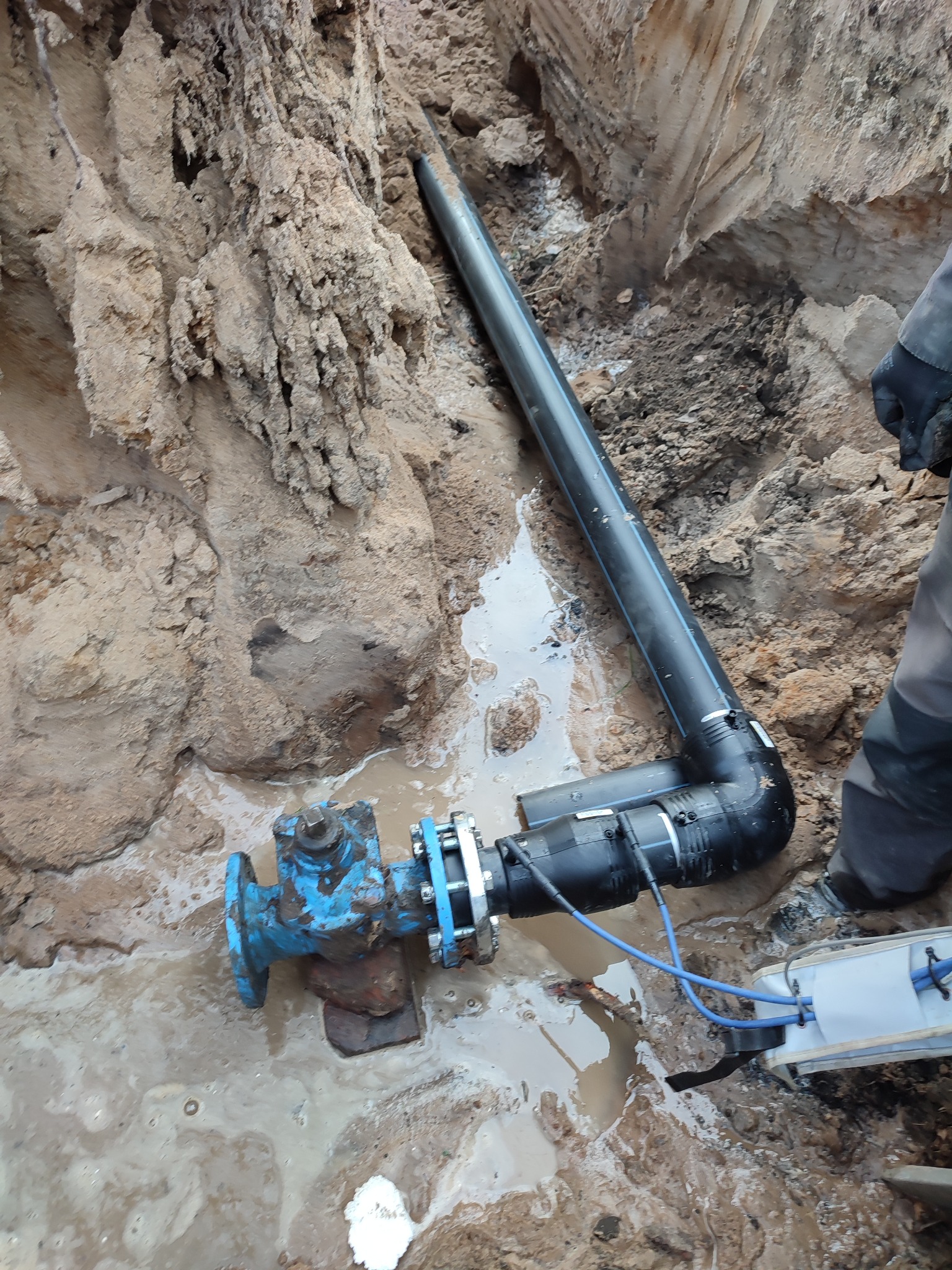 Niebieski zawór połączony z czarną rurą w wykopie wypełnionym wodą, widoczne ślady ziemi na zaworze, obok fragment narzędzia do łączenia rur i fragment osoby w roboczym ubraniu.