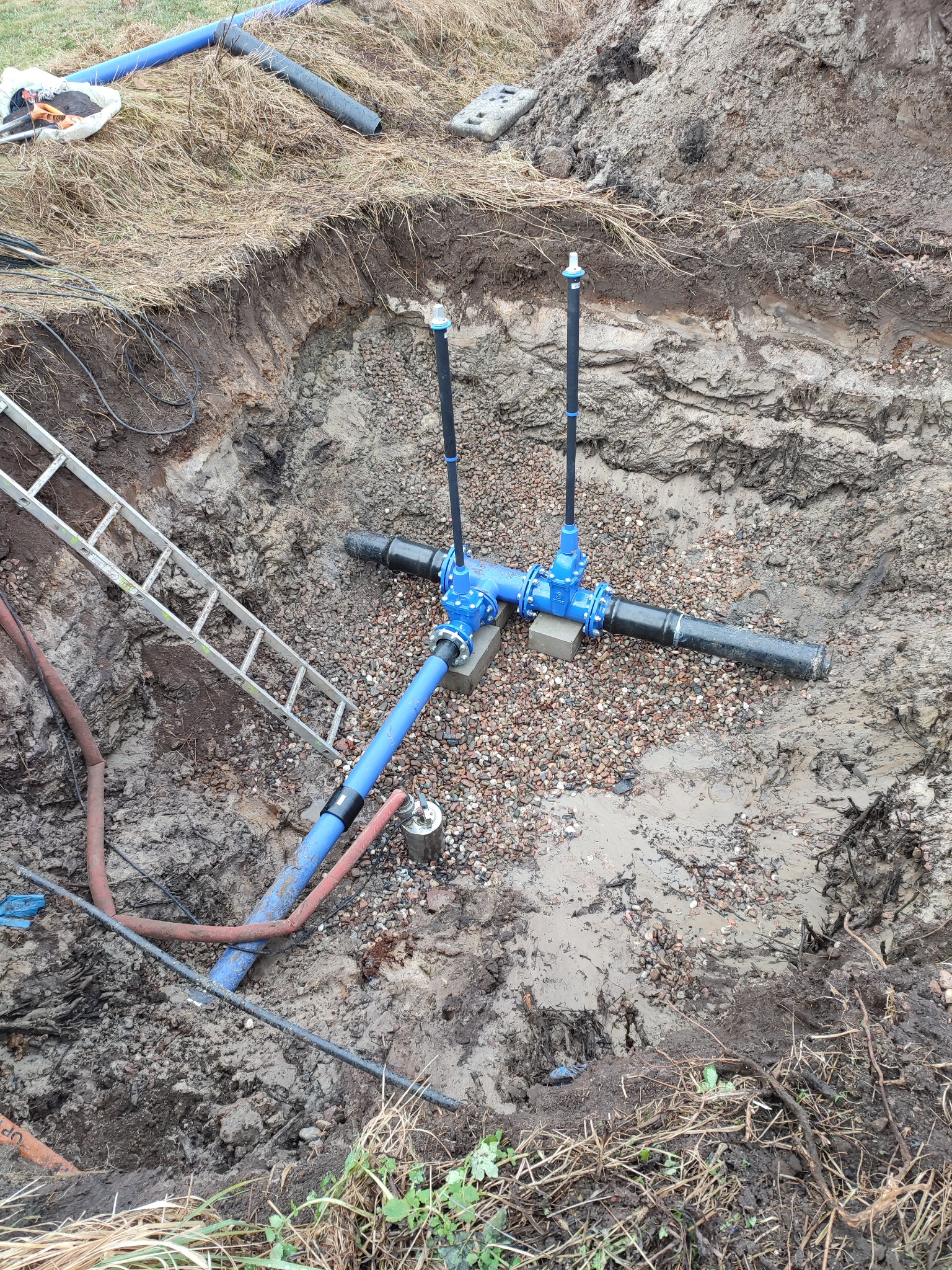 Głęboki wykop z widoczną instalacją wodociągową: niebieskie i czarne rury połączone zaworami, częściowo zasypane żwirem, drabina oparta o ścianę wykopu, ziemia wokół wykopu.